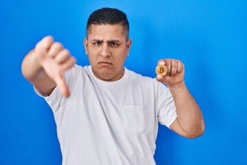 Canvas Print - Hispanic young man holding virtual currency bitcoin with angry face, negative sign showing dislike with thumbs down, rejection concept