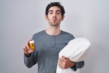 Poster - Young hispanic man wearing pyjama hugging pillow holding pills looking at the camera blowing a kiss being lovely and sexy. love expression.