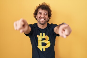 Canvas Print - Hispanic young man wearing bitcoin t shirt pointing to you and the camera with fingers, smiling positive and cheerful