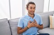 © Krakenimages.com - Young hispanic man sitting on sofa with hands on heart at home