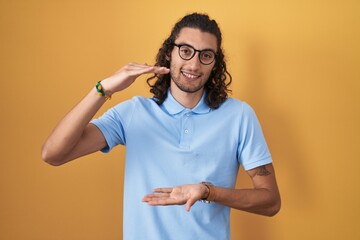 Poster - Young hispanic man standing over yellow background gesturing with hands showing big and large size sign, measure symbol. smiling looking at the camera. measuring concept.