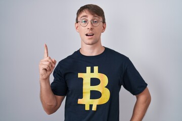 Poster - Caucasian blond man wearing bitcoin t shirt pointing finger up with successful idea. exited and happy. number one.