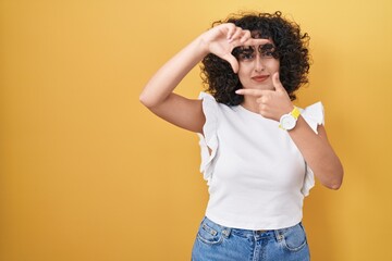 Sticker - Young middle east woman standing over yellow background smiling making frame with hands and fingers with happy face. creativity and photography concept.