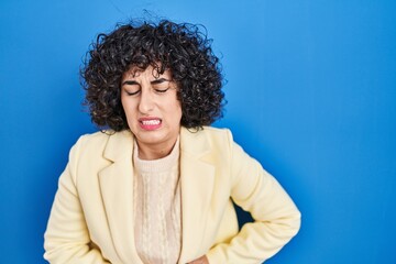 Poster - Young brunette woman with curly hair standing over blue background with hand on stomach because indigestion, painful illness feeling unwell. ache concept.