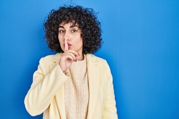 Poster - Young brunette woman with curly hair standing over blue background asking to be quiet with finger on lips. silence and secret concept.