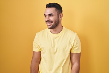 Poster - Young hispanic man standing over yellow background looking away to side with smile on face, natural expression. laughing confident.