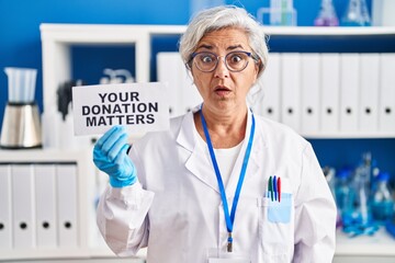 Canvas Print - Middle age woman with grey hair working at scientist laboratory holding your donation matters banner scared and amazed with open mouth for surprise, disbelief face