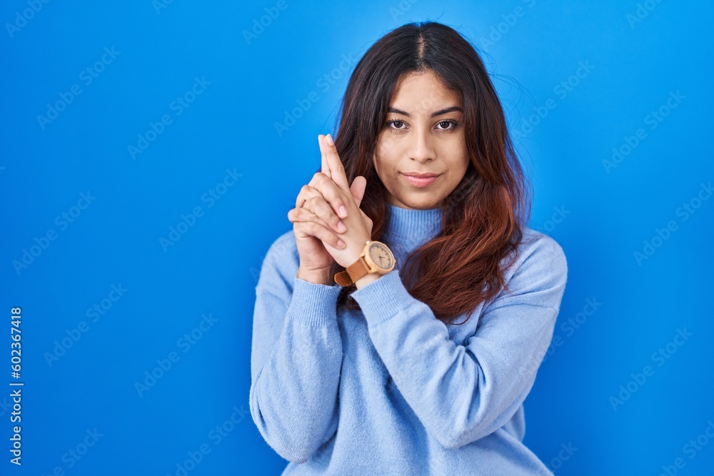 Hispanic young woman standing over blue background holding symbolic gun with hand gesture, playing killing shooting weapons, angry face