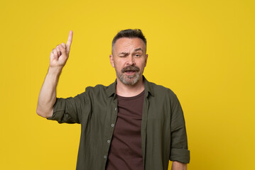 Mature 50s handsome man pointing with finger up above copy space doubtgul grimace on his face with raised eyebrow dressed casual green shirt standing over isolated yellow background