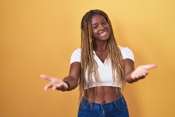 Sticker - African american woman with braided hair standing over yellow background smiling cheerful offering hands giving assistance and acceptance.