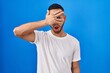 © Krakenimages.com - Young hispanic man standing over blue background peeking in shock covering face and eyes with hand, looking through fingers with embarrassed expression.