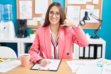 Sticker - Young hispanic woman working at the office wearing glasses smiling doing phone gesture with hand and fingers like talking on the telephone. communicating concepts.