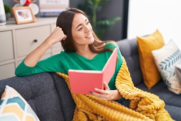 Poster - Young beautiful hispanic woman reading book sitting on sofa at home