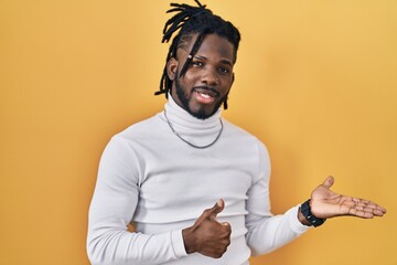 Wall Mural - African man with dreadlocks wearing turtleneck sweater over yellow background showing palm hand and doing ok gesture with thumbs up, smiling happy and cheerful