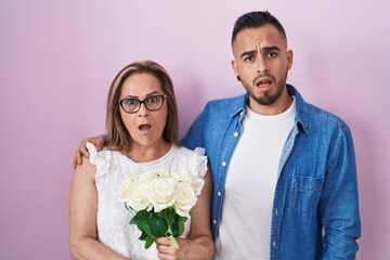 Poster - Hispanic mother and son together holding bouquet of white flowers in shock face, looking skeptical and sarcastic, surprised with open mouth