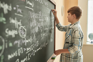 Side view portrait of young schoolboy writing on blackboard in class, copy space
