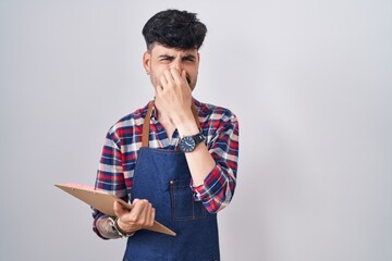 Poster - Young hispanic man with beard wearing waiter apron holding clipboard smelling something stinky and disgusting, intolerable smell, holding breath with fingers on nose. bad smell