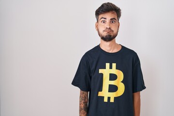 Wall Mural - Young hispanic man with tattoos wearing bitcoin t shirt puffing cheeks with funny face. mouth inflated with air, crazy expression.