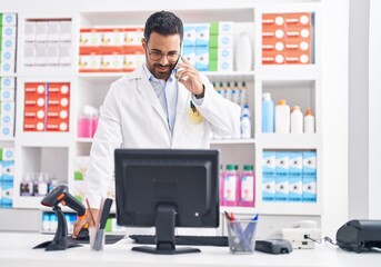 Canvas Print - Young hispanic man pharmacist talking on smartphone using computer at pharmacy