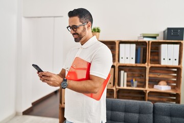 Wall Mural - Young hispanic man psychologist using smartphone holding clipboard at psychology center