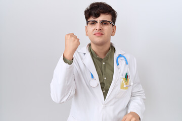 Sticker - Young non binary man wearing doctor uniform and stethoscope angry and mad raising fist frustrated and furious while shouting with anger. rage and aggressive concept.