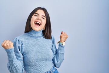 Wall Mural - Young hispanic woman standing over blue background celebrating surprised and amazed for success with arms raised and eyes closed. winner concept.