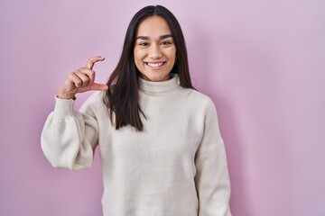 Wall Mural - Young south asian woman standing over pink background smiling and confident gesturing with hand doing small size sign with fingers looking and the camera. measure concept.