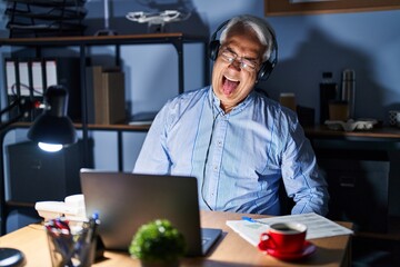 Wall Mural - Hispanic senior man wearing call center agent headset at night sticking tongue out happy with funny expression. emotion concept.