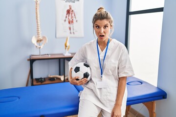 Canvas Print - Young woman working at football therapy clinic scared and amazed with open mouth for surprise, disbelief face