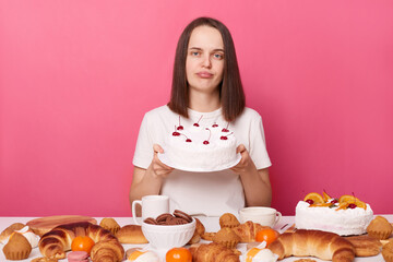 Wall Mural - Sad unhappy woman wearing white T-shirt sitting at festive table with various desserts, isolated over pink background looking at camera with pout lips holding cake.