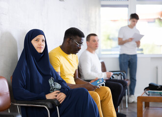 Wall Mural - Young arab woman and adult african man in casual clothes waiting for their turn on chair at reception