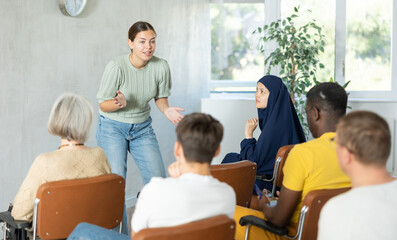 Wall Mural - Emotional young female tutor leading educational class for multiethnic adult group sitting in auditorium