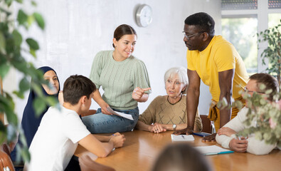 Wall Mural - Smiling young girl participating in volunteer work discussing goals and objectives of project or event with group of associates, men and women of different ages and nationalities gathered around table