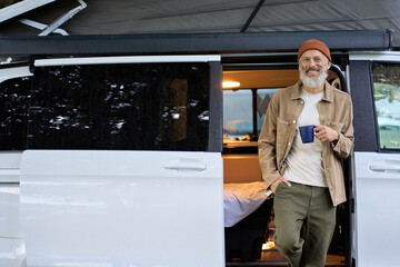 Wall Mural - Happy older senior hipster man standing near rv camper van on vacation. Mature traveler looking at camera, holding drinking coffee waking up in the morning in camping tourism nature park.