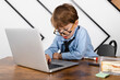© Georgi - Little boy in round eyeglasses, blue shirt and tie sitting at the desk in the office, working in the laptop. Smart little kid, Child prodigy.