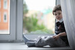 © alexkich - The little boy is reading a book. The child sits at the window and prepares for lessons. A boy with a book in his hands is sitting on the windowsill.