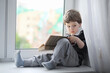 © alexkich - The little boy is reading a book. The child sits at the window and prepares for lessons. A boy with a book in his hands is sitting on the windowsill.