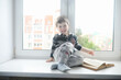 © alexkich - The little boy is reading a book. The child sits at the window and prepares for lessons. A boy with a book in his hands is sitting on the windowsill.
