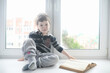 © alexkich - The little boy is reading a book. The child sits at the window and prepares for lessons. A boy with a book in his hands is sitting on the windowsill.