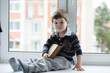 © alexkich - The little boy is reading a book. The child sits at the window and prepares for lessons. A boy with a book in his hands is sitting on the windowsill.