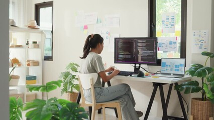 Side view of professional young Asia girl IT development programmer typing on keyboard coding programming fixing data code on computer screen and laptop on table in workroom at house office.