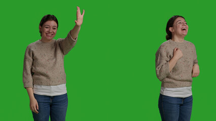 Wall Mural - Close up of female model greeting someone and waving, smiling in studio. Relaxed young woman waving hi or hello on camera, standing over full body greenscreen backdrop and saying goodbye.