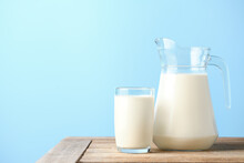 Glass And Pitcher Of Fresh Milk On Wooden Table With Light Blue Background.