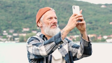 Wall Mural - Cool bearded happy older elder hipster man standing in nature park taking photos on smartphone using mobile phone. Mature boomer traveler enjoying vacation trip making landscape or selfie picture.