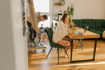 Woman working on laptop by man and daughter loading utensils in dishwasher