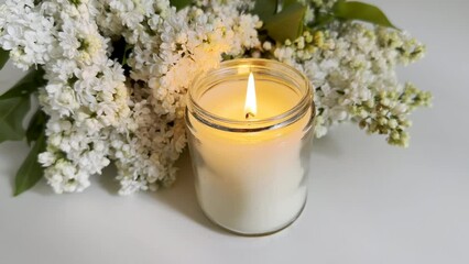 Wall Mural - Burning candle in clear glass jar and white lilac bouquet on white table, candle video mockup