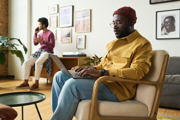 Wall Mural - Young serious African American analyst typing on laptop keyboard while sitting in armchair against male coworker talking on smartphone