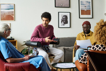 Leinwandbilder - Young multi-ethnic economist explaining something to mature female coworker while sitting in circle of intercultural colleagues at meeting