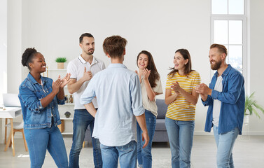 Joyful enthusiastic friends applaud man expressing their respect and congratulating him on victory. Young multiracial people clapping hands to male friend while standing in living room at home.