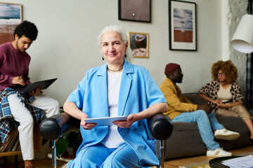 Poster - Happy mature grey haired female chief executive officer with tablet sitting in armchair in front of camera against youn intercultural subordinates